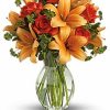 Orange roses and peach lilies arranged with greenery in a clear glass vase.